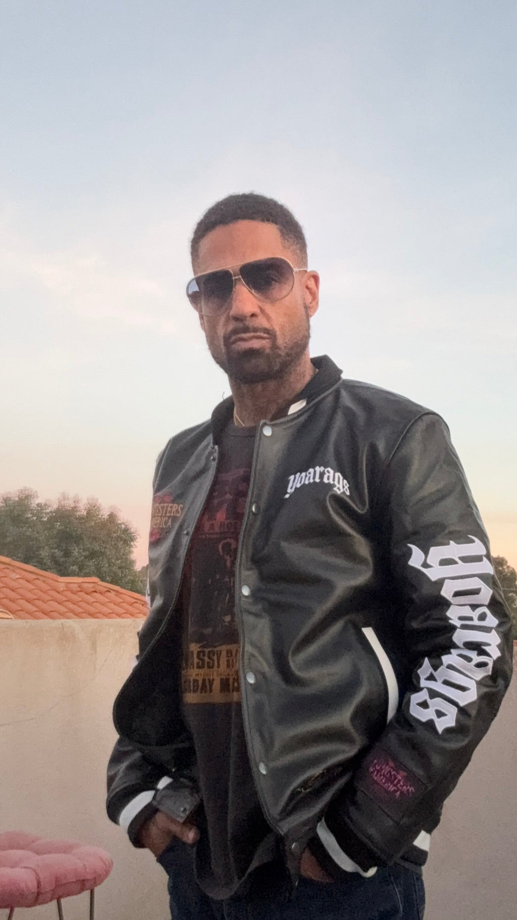 A man wearing a black YOA LEATHERS varsity jacket with 'YOARAGS' in white Gothic lettering on the sleeves. dark jeans, and sunglasses, standing outdoors on a patio.